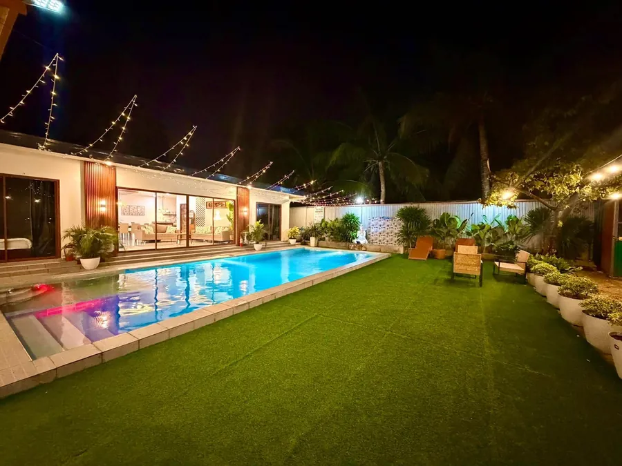 Long private pool beside the lit villa, with string lights and a grass lawn at night
